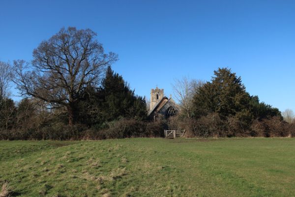 A view over an open field to Rampton Church, bathed in blue skies and surrounded by trees